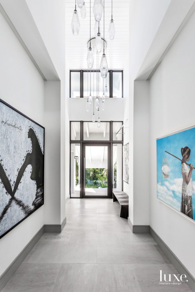 A glass WSI pivot door opens to the foyer of a renovated Coral Gables home by designer Carolyn McCarthy. The drama continues with Hammerton Studio's Aalto pendants as well as La Isla Aquella by Jose Bedia (left) and Dreamcatcher by Sandro de la Rosa (right). Artefacto's Sweep bench rests on leathered marble flooring from Innovative Surfaces.