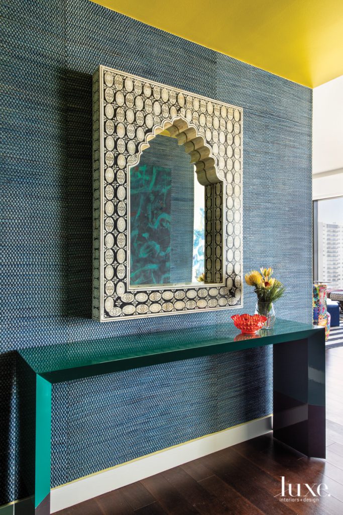 The entry to this Houston high- rise introduces the vivid color palette throughout. A Phillip Jeffries wallcovering frames a vintage black-and-white inlay mirror from Carl Moore Antiques over a custom table with a dual lacquered finish. Art visible in the reflection by Tony Magar was selected by art consultant Maura Parro from Laura Rathe Fine Art.