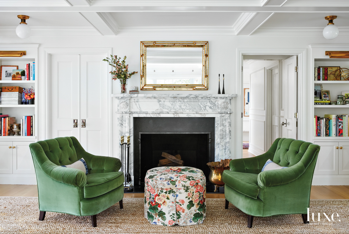 A seating area in front of the living room fireplace with green velvet chairs, a floral ottoman and a vintage mirror.