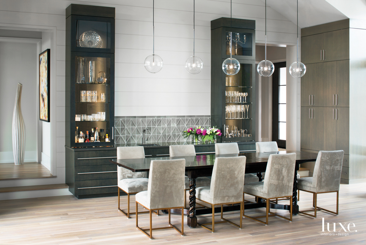 A dining room has a long table, upholstered chairs and a row of glass pendants above. Built-in cabinets line one wall.