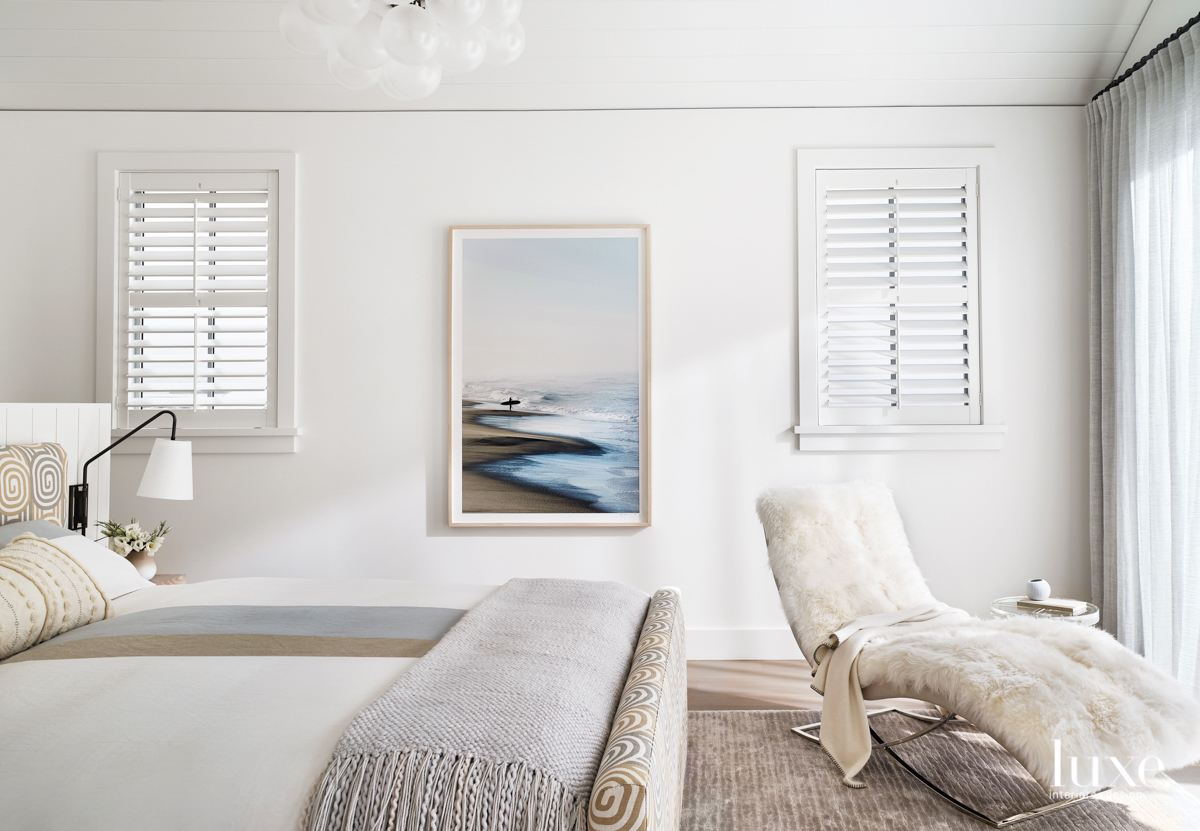 A gorgeous photograph of the shoreline sits between two shuttered windows in this cream-colored, minimalistic master bedroom.