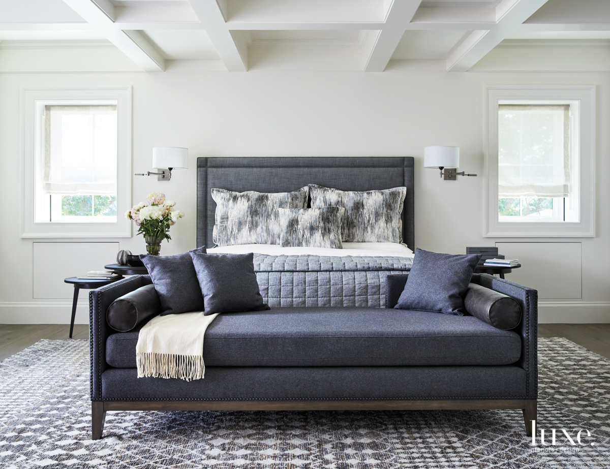 A master bedroom is done in blue and gray. A plush bench sits at the end of the bed.