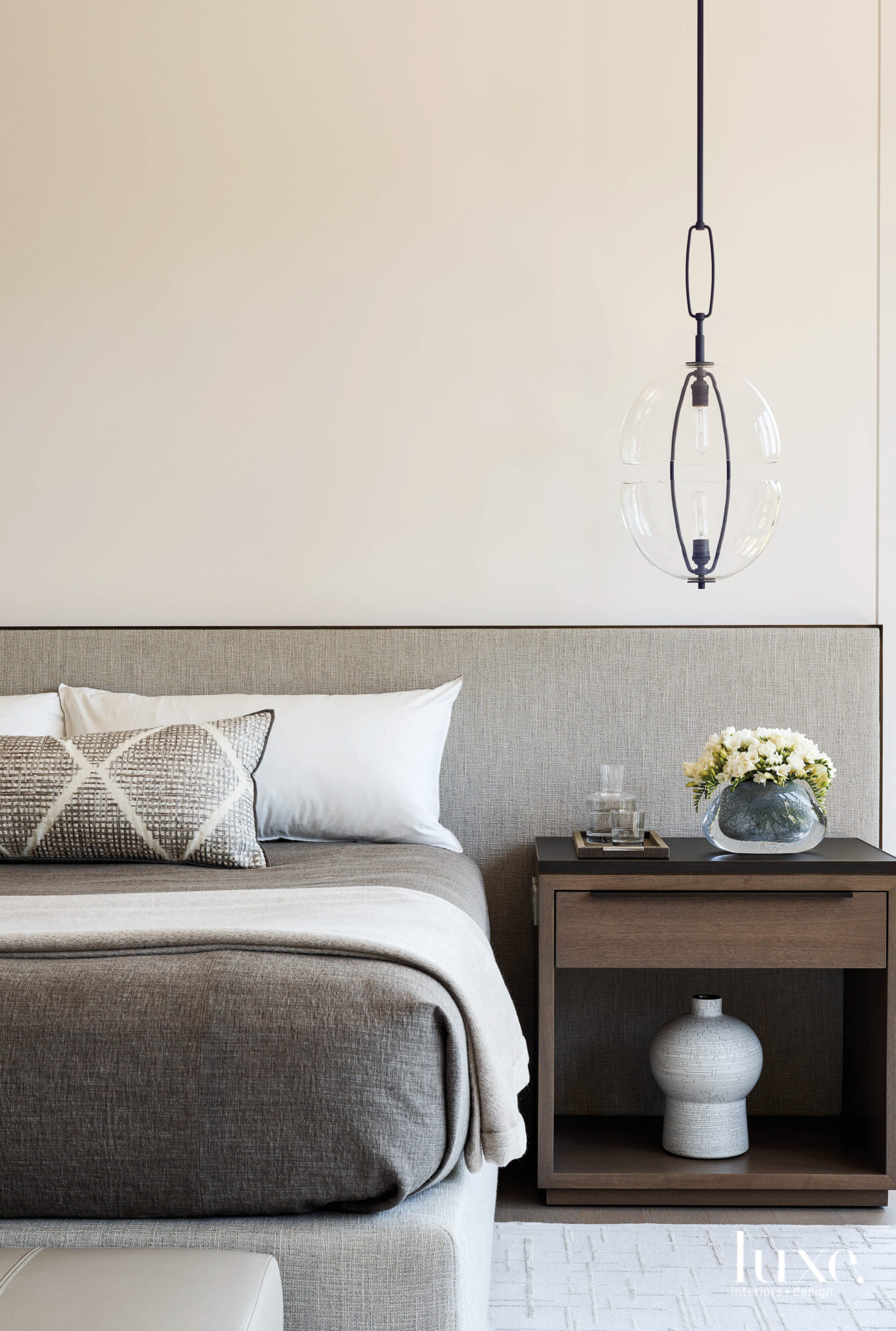 A bedroom has a long upholstered headboard and neutral linens.