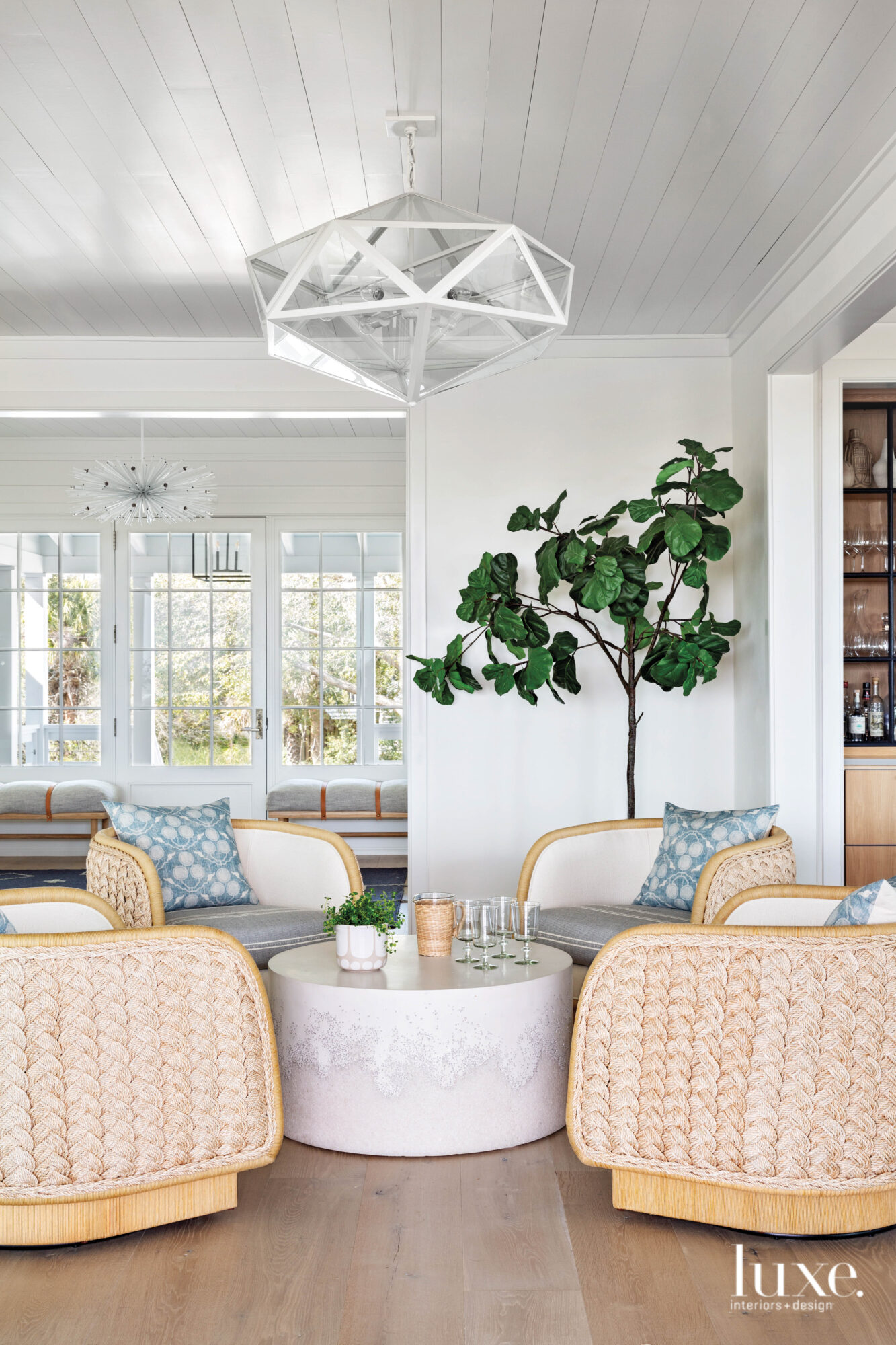 Sitting area with four chairs, drum cocktail table, fiddle leaf fig tree, pendant, bank of windows beyond