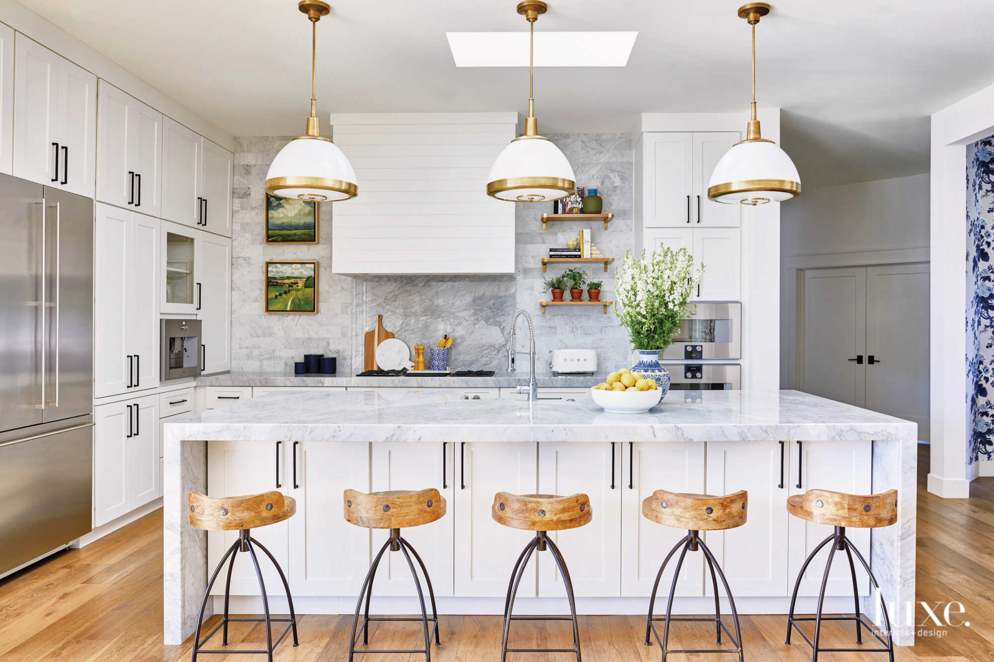 The white kitchen has white and gold pendant lighting, marble countertops and wooden stools.