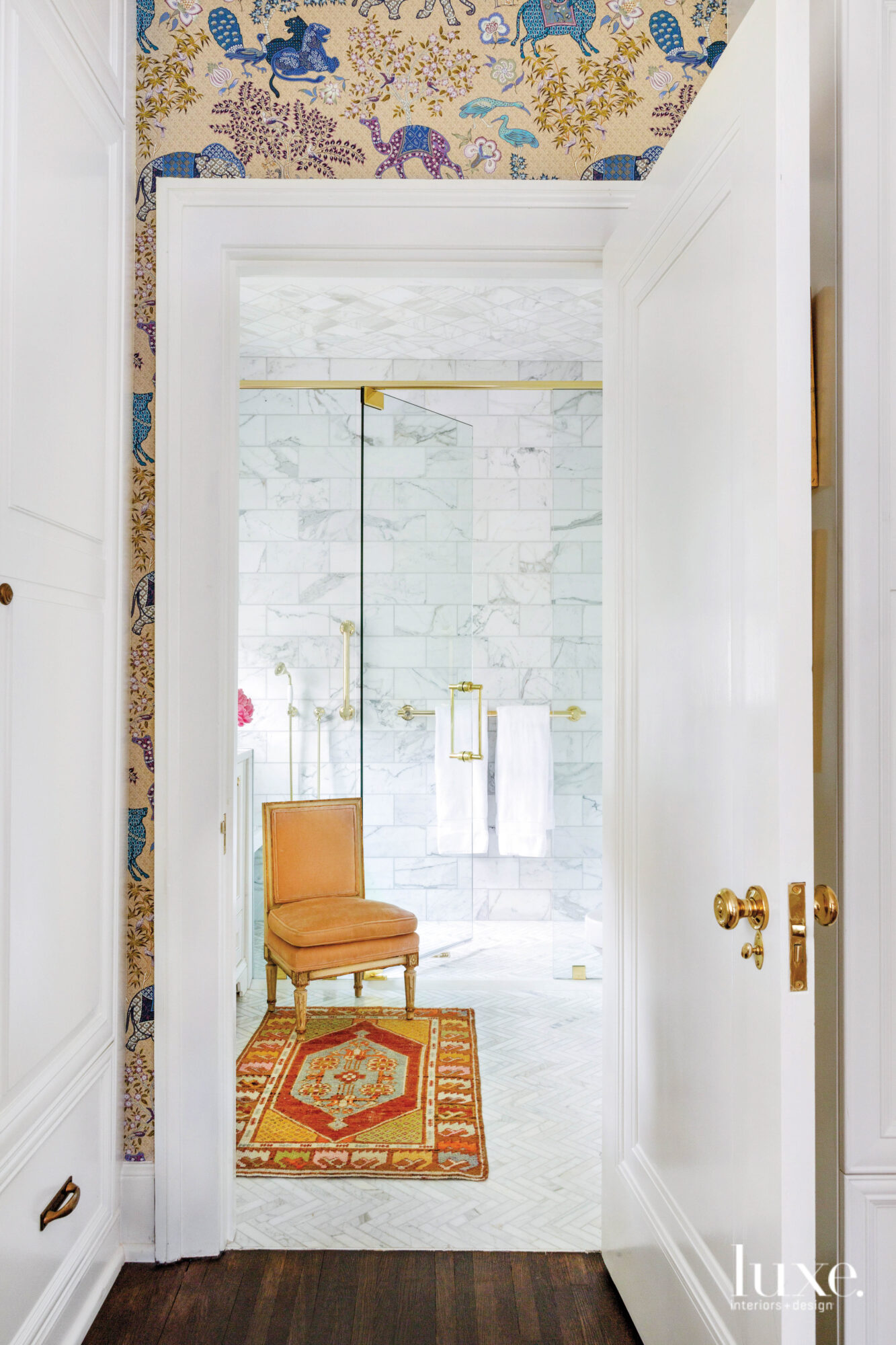Bedroom with white marble, clear glass shower, brass hardware, an orange chair and an orange rug