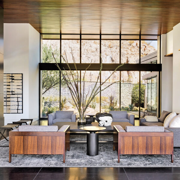A living room with a floor-to-ceiling stone fireplace, gray and wood furnishings, and a granite floor. Floor-to-ceiling windows look out on a desert garden.