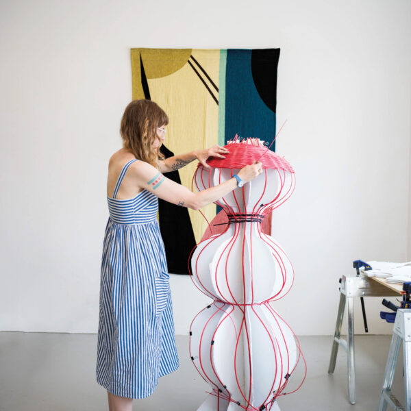 The artist weaving a large pink basket. A textile hangs on the wall.