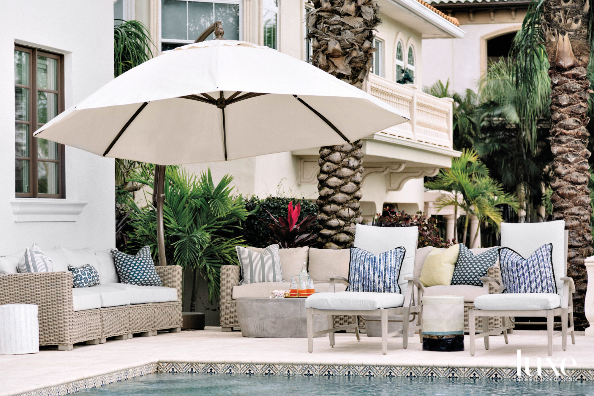 Chaises and sofas shaded by a large umbrella and next to a pool.