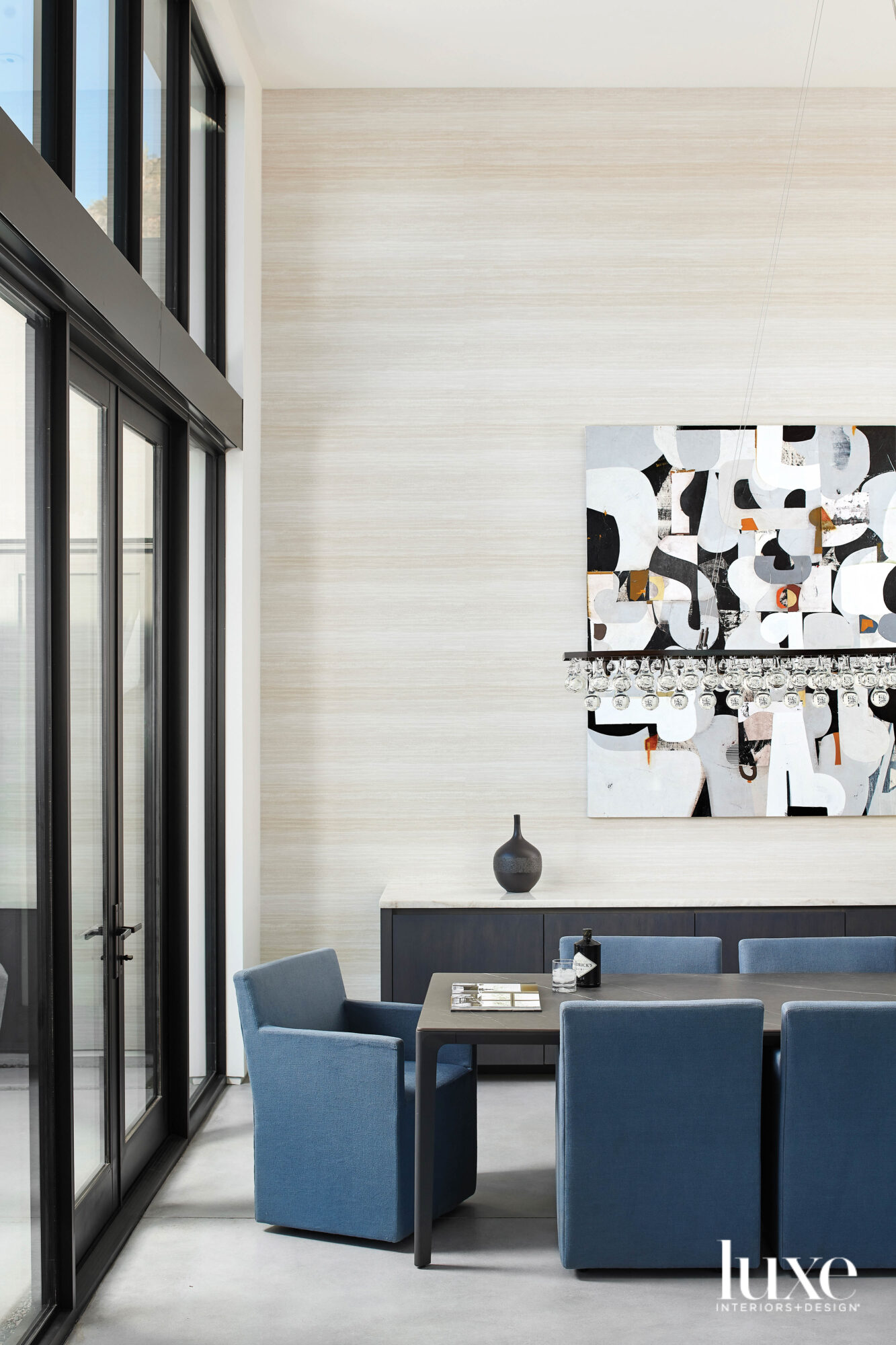 In the dining room, an abstract painting hangs behind a large black dining table surrounded by blue upholstered chairs.