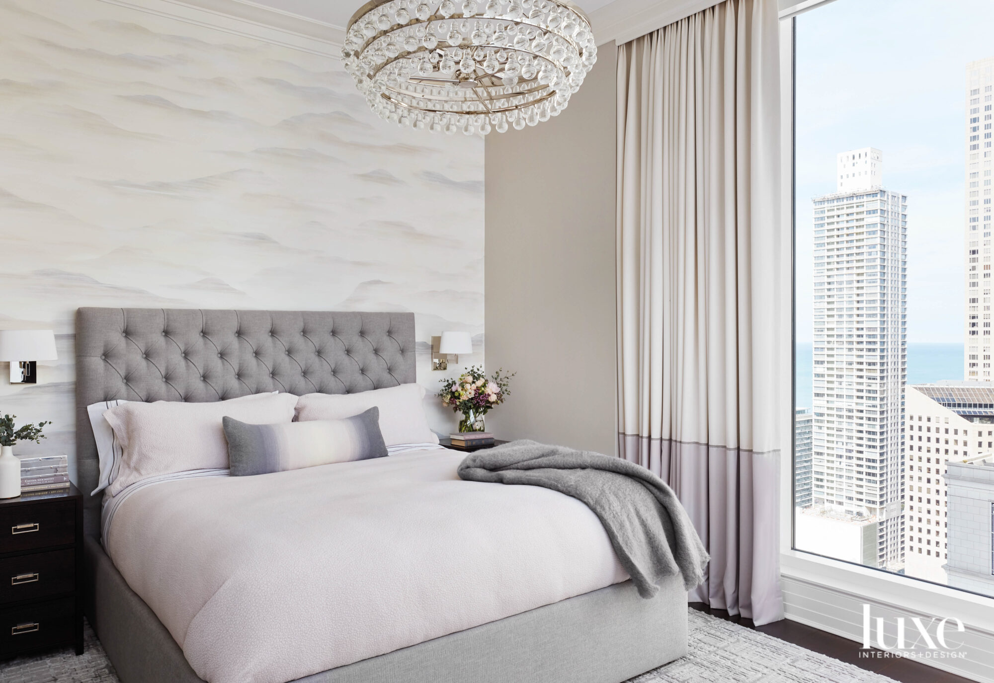 A bedroom with a gray bed, cloud wallpaper and a crystal chandelier. It looks out on the city of Chicago.