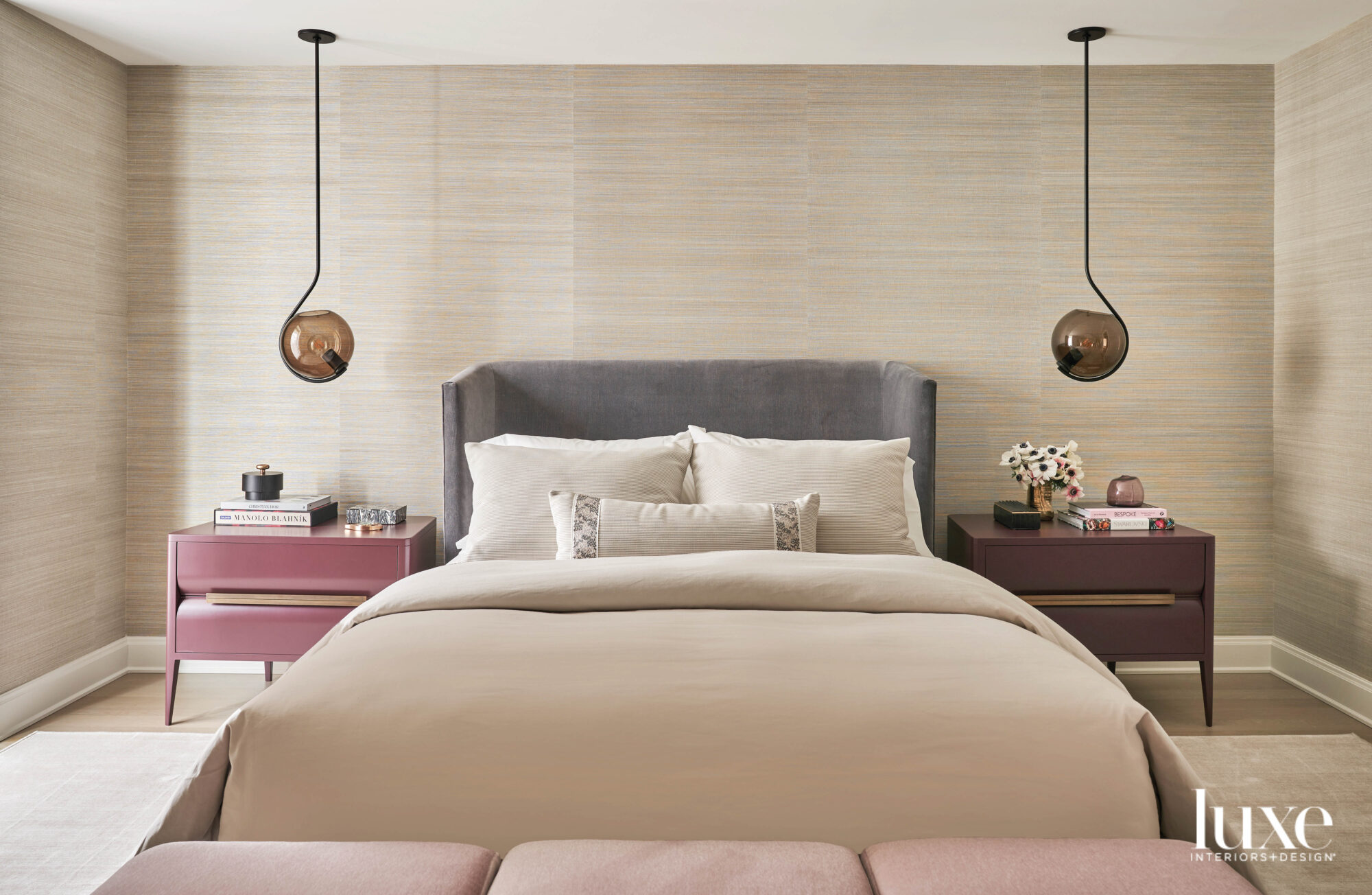 A bedroom with tan striped wallpaper, pink nightstands and a gray bed with tan linens.