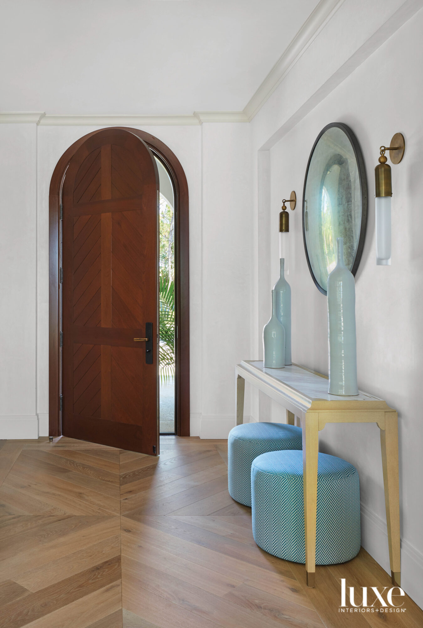 entry with chevron pattern flooring, curved wood door, blue stools, wood console and round mirror