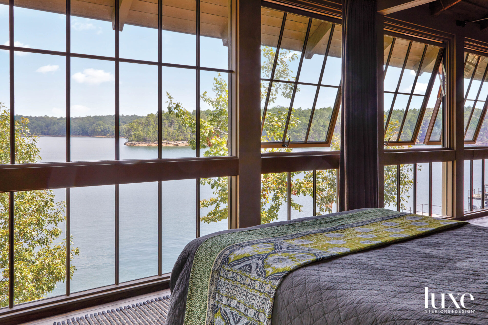 Edge of a bed looking out onto a bank of floor-to-ceiling windows with tilt-out casements revealing views of a placid lake