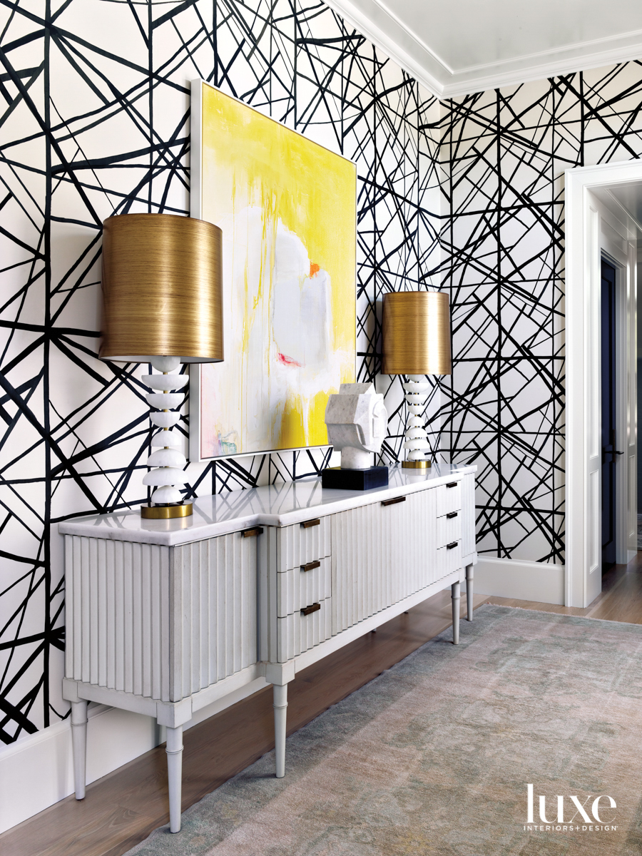 Sideboard topped with a pair of table lamps wearing brass shades, a large abstract yellow canvas, and a backdrop of dramatic black-and-white wallpaper