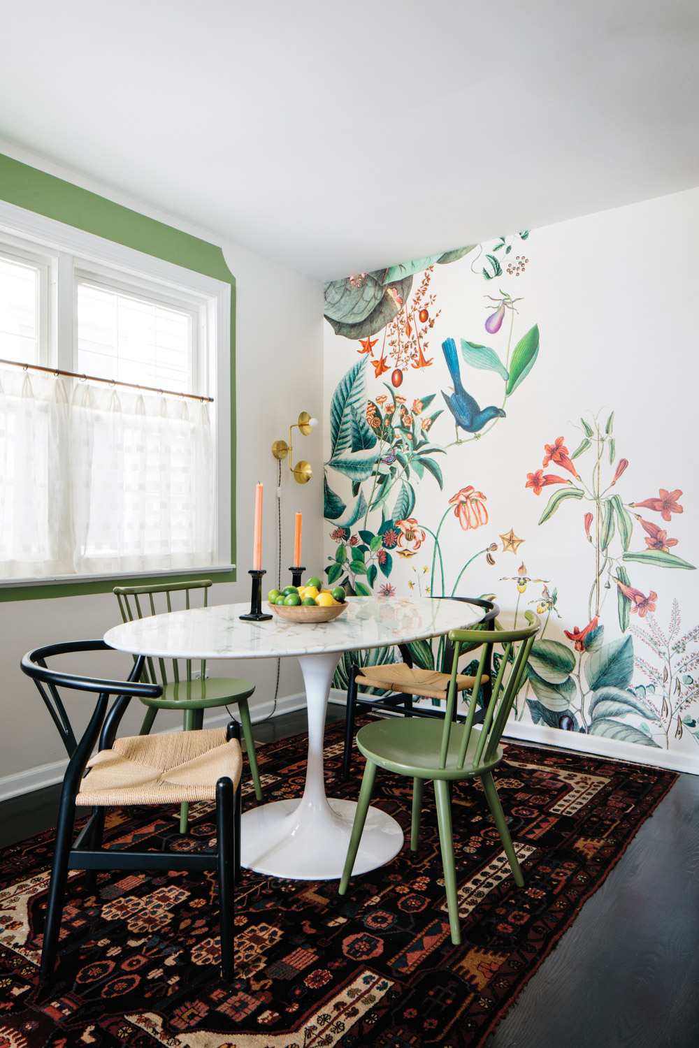 Dining area with white table, green and black chairs and a tropical-print wallcovering.