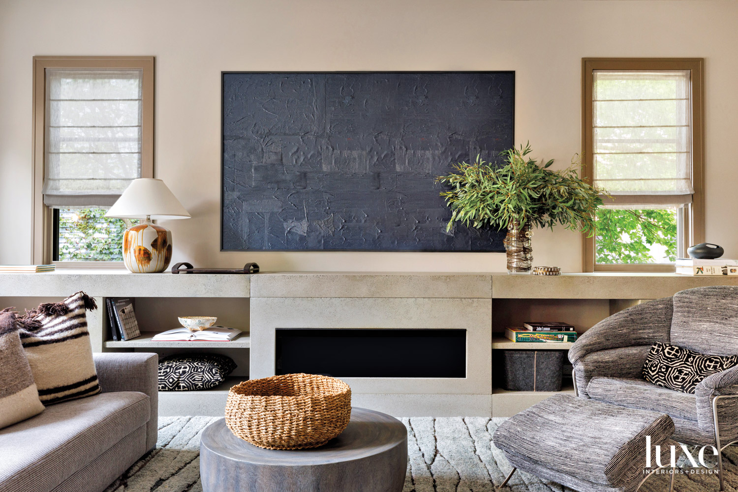 In a neutral living room a black, textured painting hangs above a concrete fireplace.