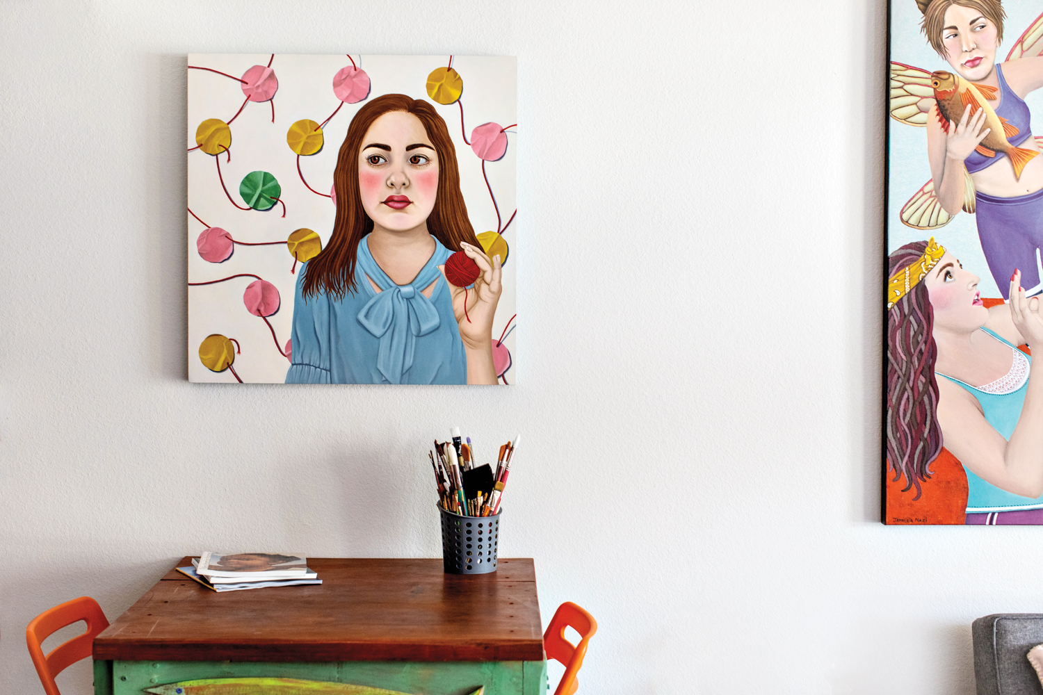 A painting of a girl in a blue shirt holding a red ball of yarn. Below is a desk with paint brushes on top. To the right, you can see part of another painting.