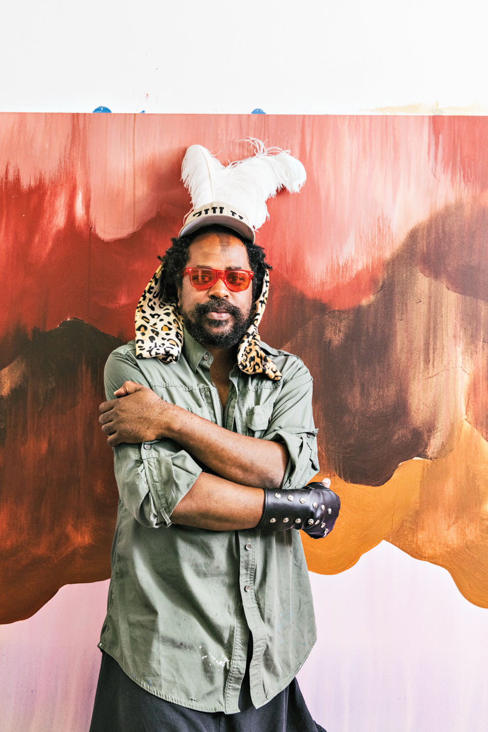 artist umar Rashid poses against a painted wall wearing red sunglasses and a hat with white feathers