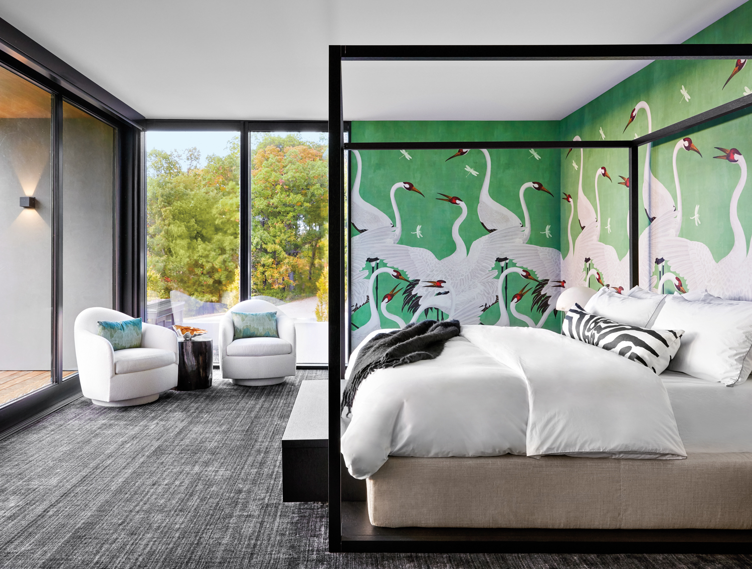 A bedroom with a dramatic green wallcovering with white herons. There is a black four-poster bed and two white swivel chairs.