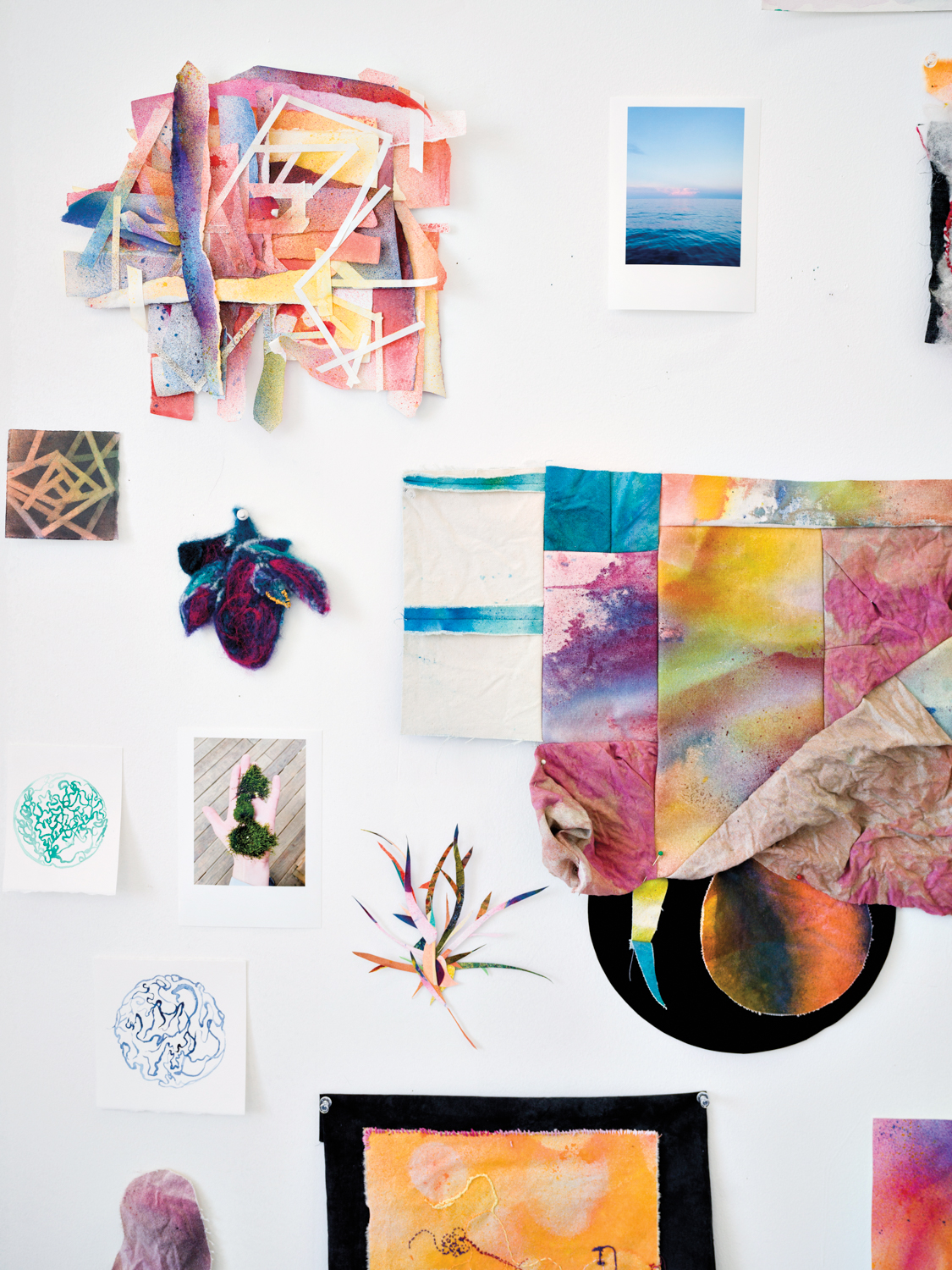 A studio wall with colorful artworks on paper and fabric, and photos from the environment.