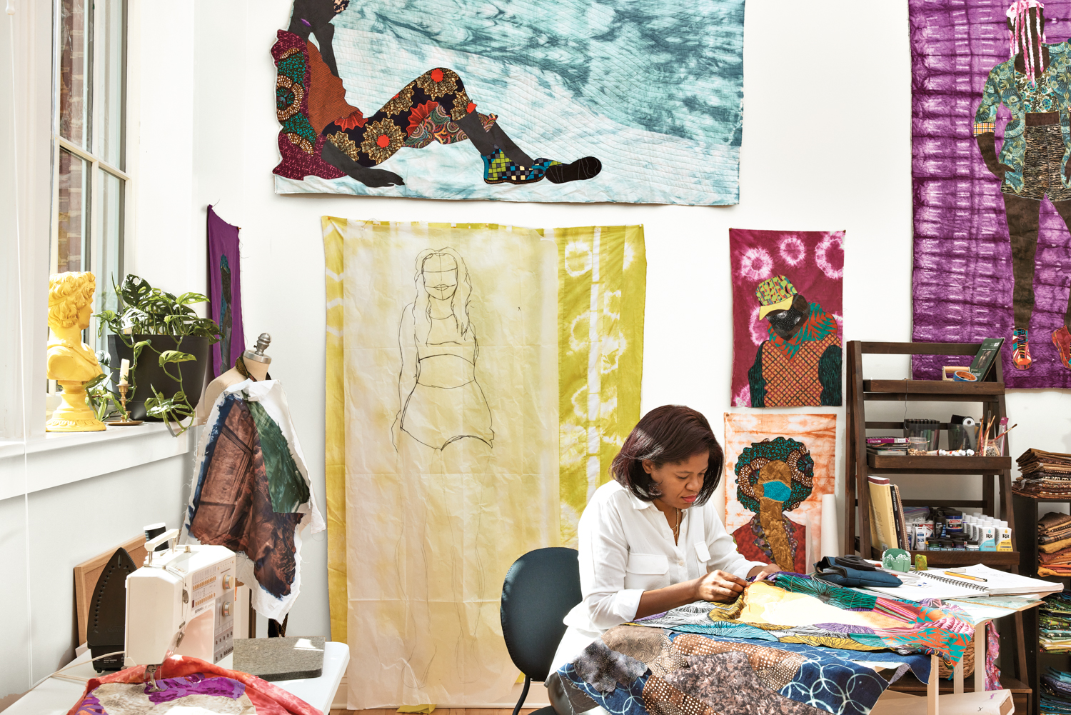 Woman hand-sewing at a desk in a room surrounded by artworks
