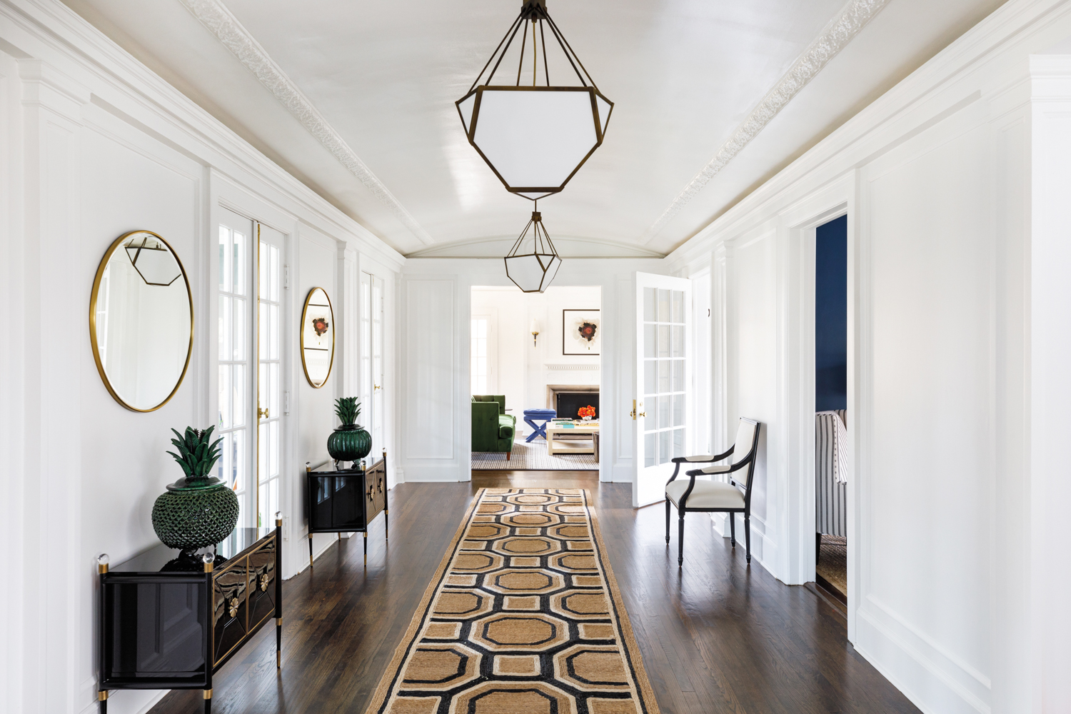 A white hallway with a cove ceiling and plaster molding details. A tan-and-black geometric rug runs the length of the hallway. Oversize geometric pendants hang from above.