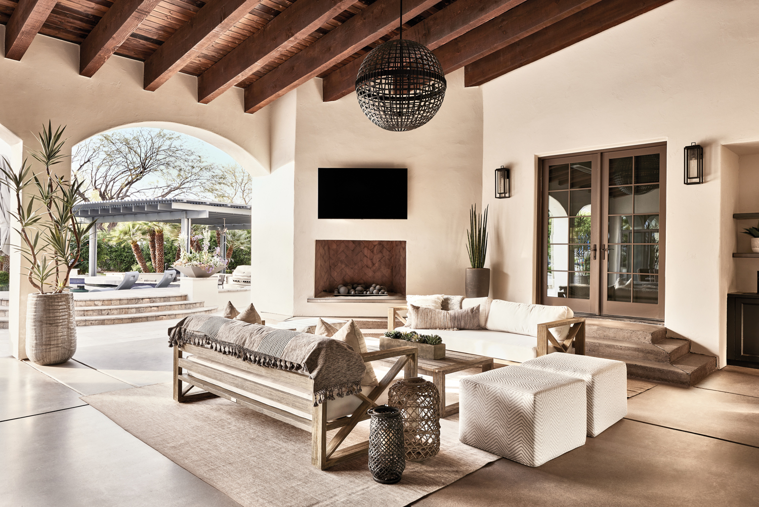 A patio with a television over the fireplace. Two wood-framed sofas upholstered in ivory fabric face each other with a wood coffee table in beetween. Two herringbone-patterned stools sit at the end.