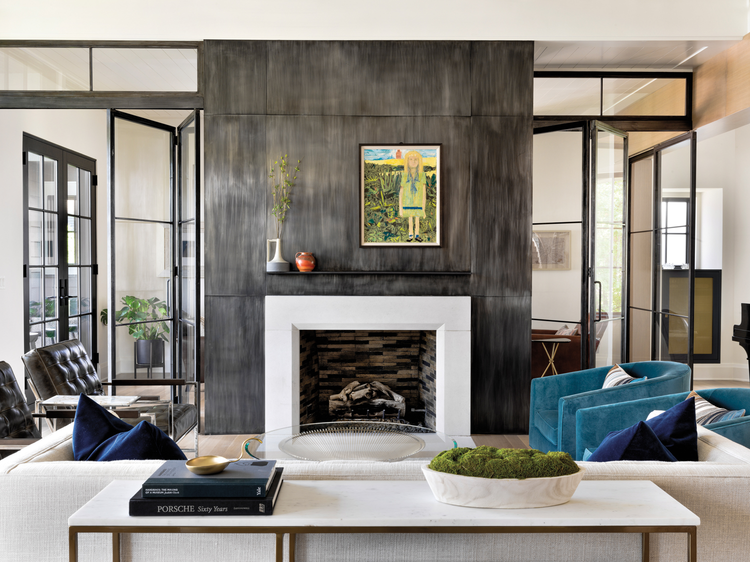 A fireplace with a steel surround is the focal point of a living room. It is surrounded by a white couch, two blue swivel chairs and two leather armchairs. A painting of a young girl hangs above the fireplace.