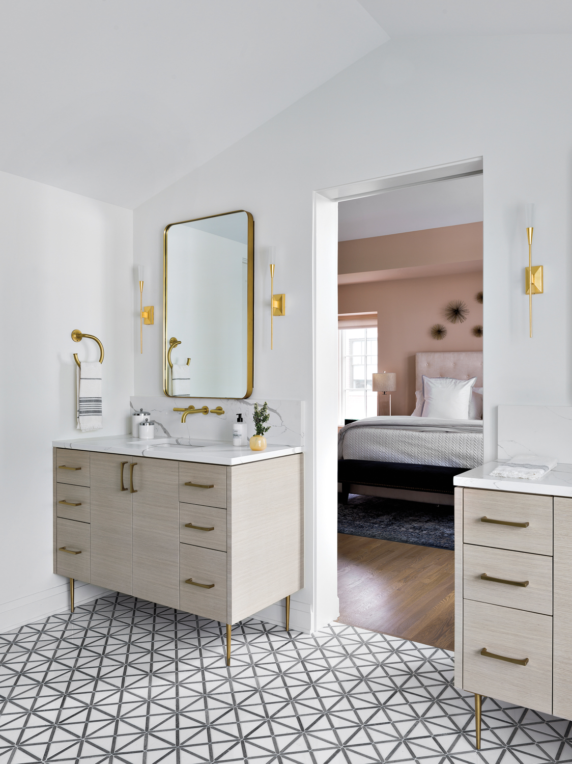 A bathroom with a geometric tile floor and matching white oak vanities. It provides a peek into the main bedroom which has pink walls.