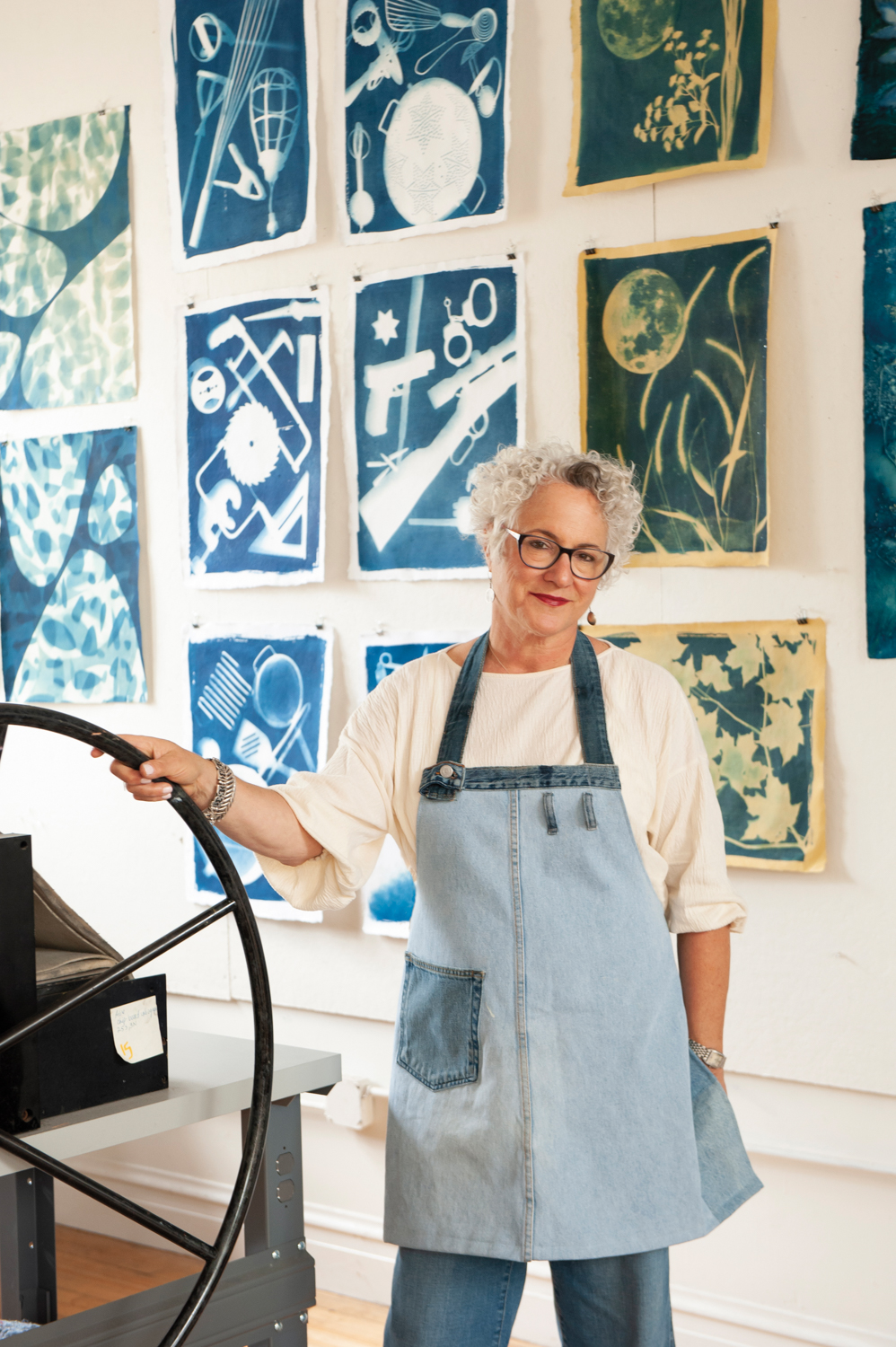 A white woman with white hair in a denim apron stands in front of blue cyanotypes.