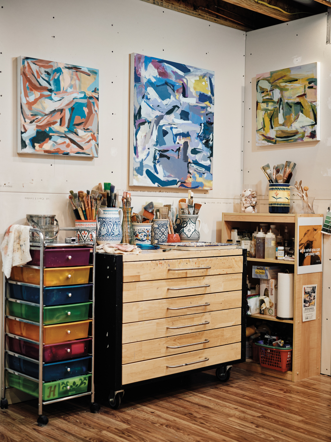 storage cabinets and shelves filled with painting supplies, with abstract paintings hung above