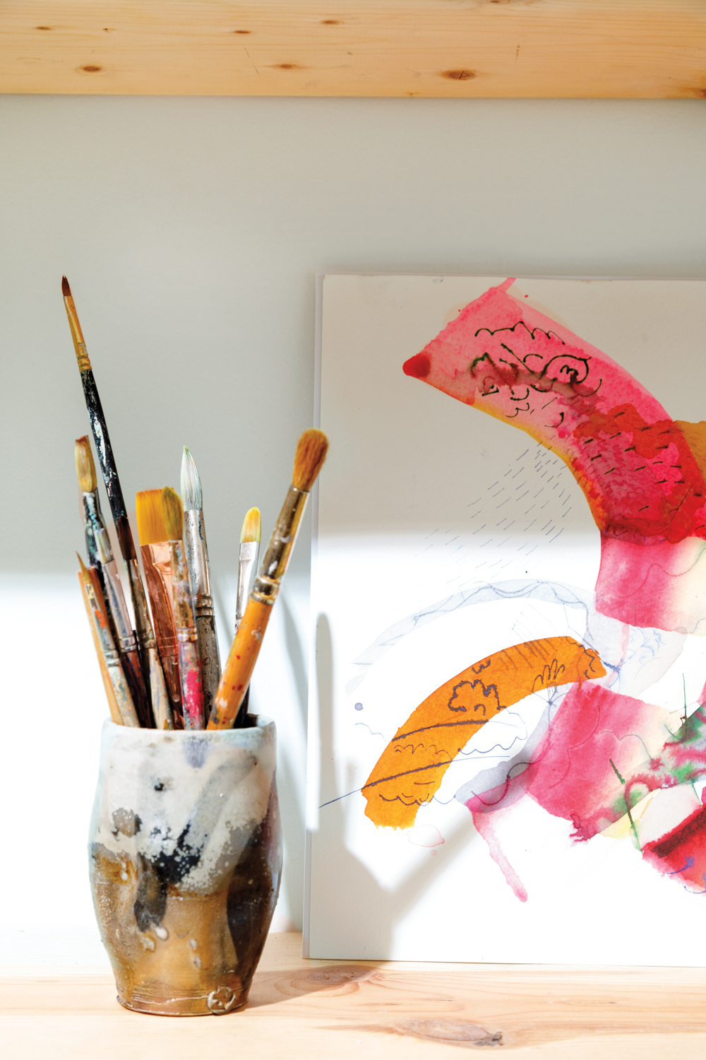 Decorative ceramic cup containing paint brushes beside abstract watercolor painting