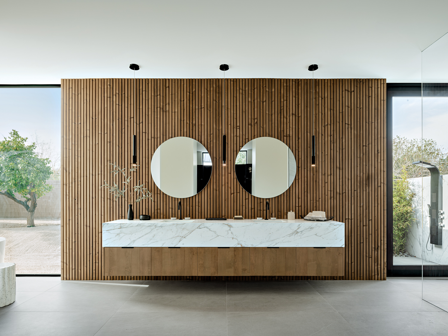 A main bathroom with wood-slatted walls, a floating marble vanity and two round mirrors.