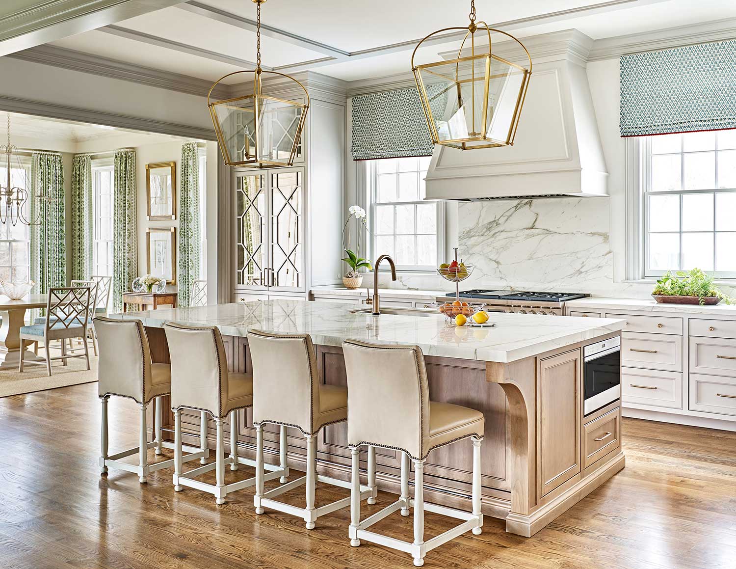 kitchen with white cabinetry and large range hood, green patterned roman shades, marble countertops and backsplash and tan counter stools
