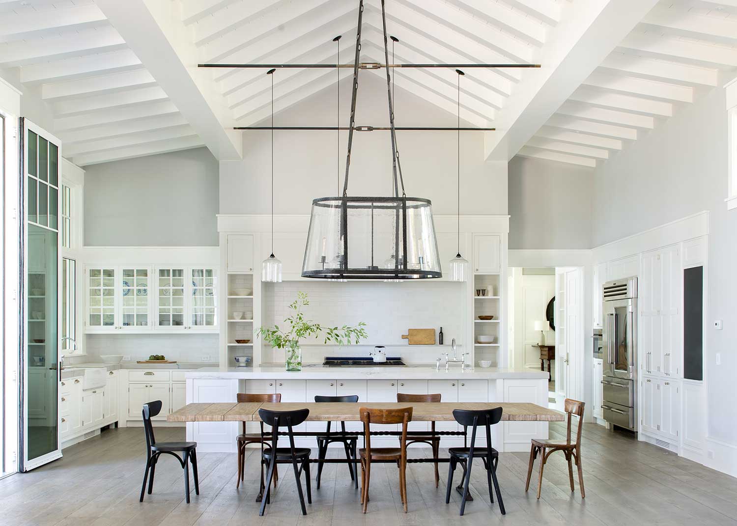 white kitchen with beams on tall ceiling, black iron lighting pendants and long wood dining table and chairs