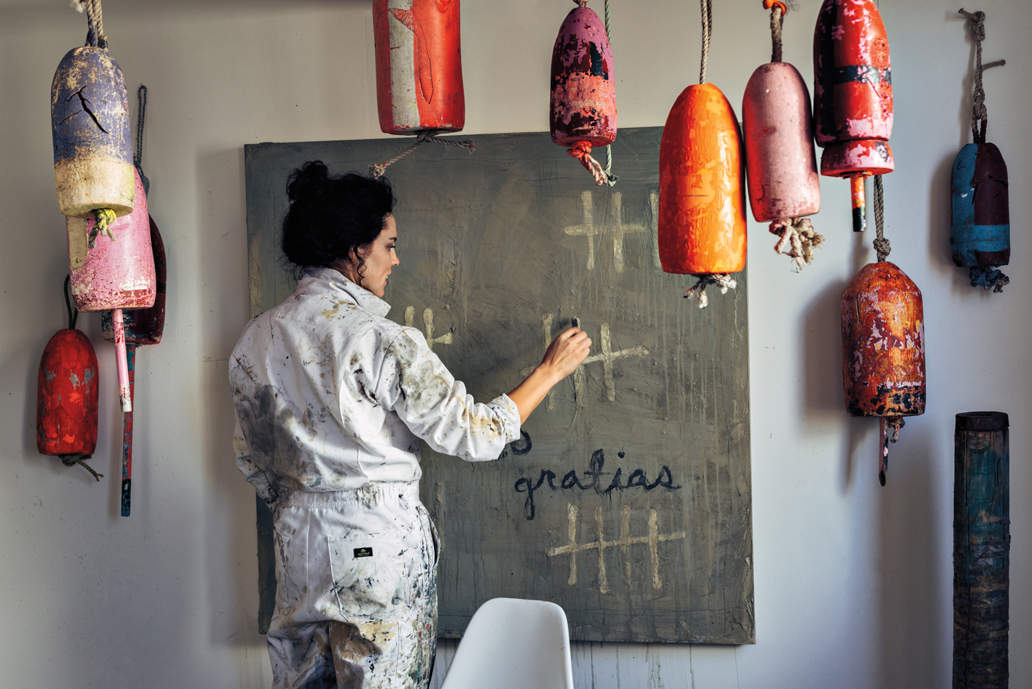 Woman standing before a canvas with tools; brightly colored objects dangle from the ceiling in the foreground
