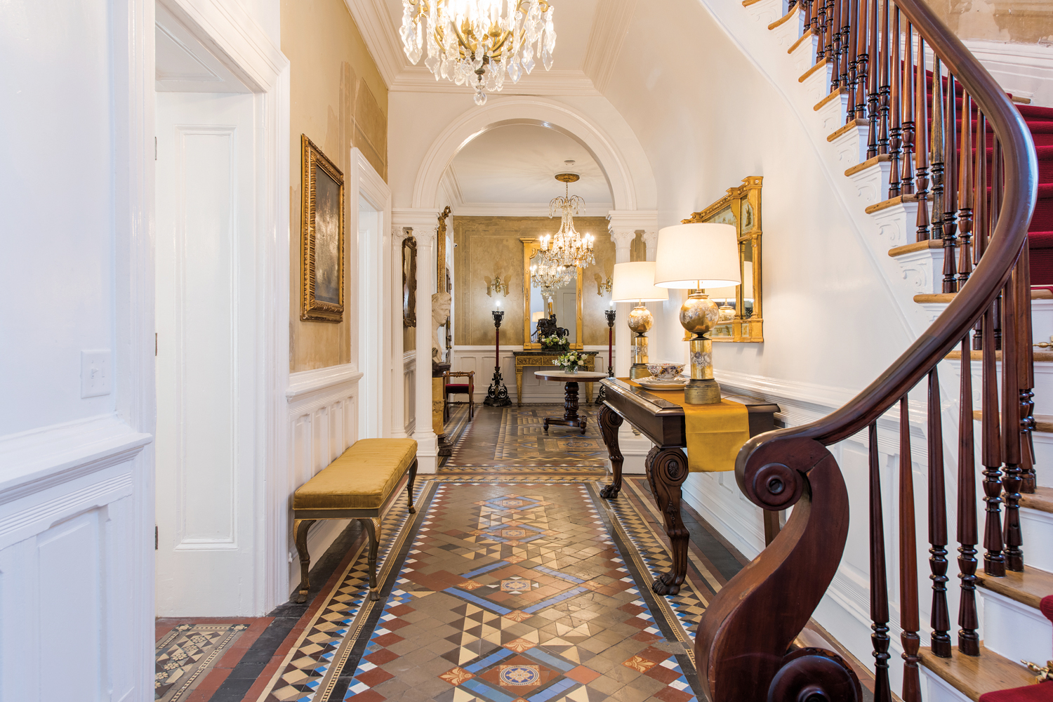 Entryway with crystal chandeliers and tiled floors; on the right, a wooden staircase leads up with a red stair runner 