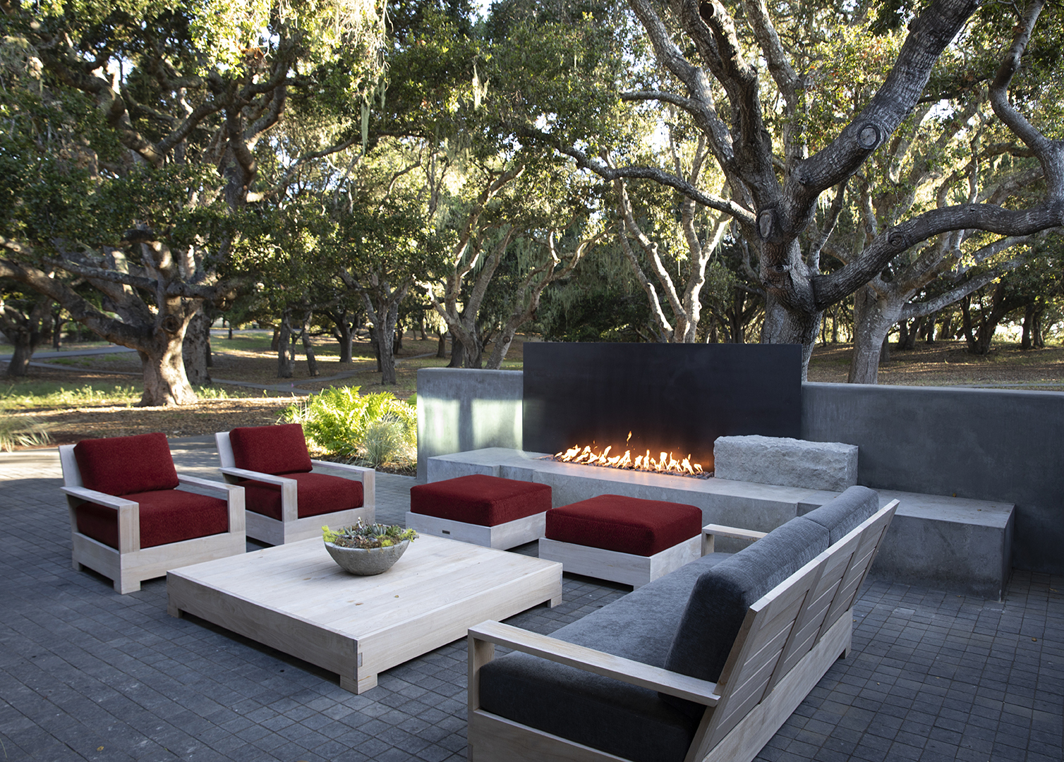 A bosque of trees providing natural shade to an outdoor seating group with red upholstery and an accompanying fire feature
