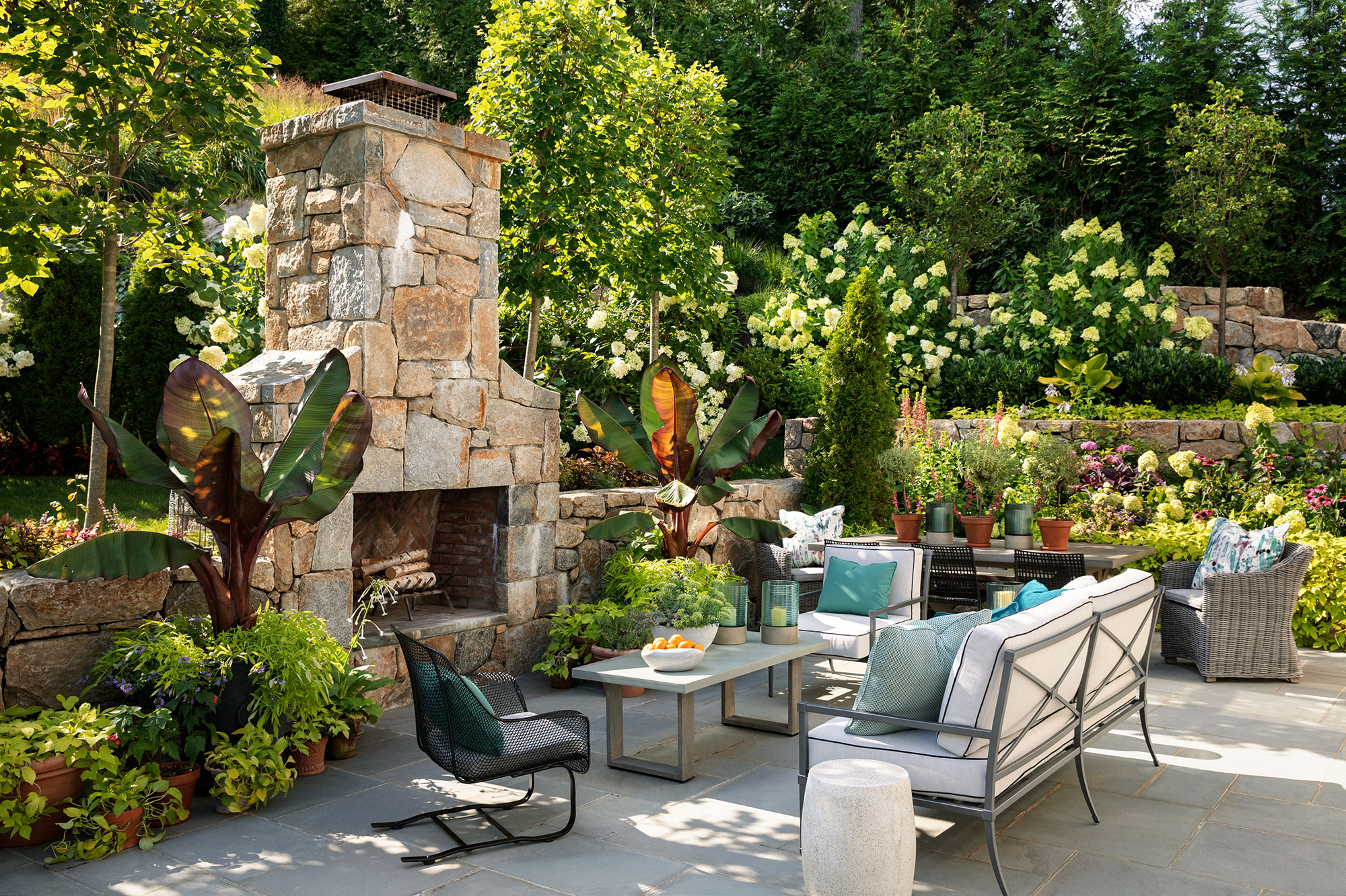 Colorful, inviting outdoor seating group in front of a stone fireplace and beside a large table topped with potted plants