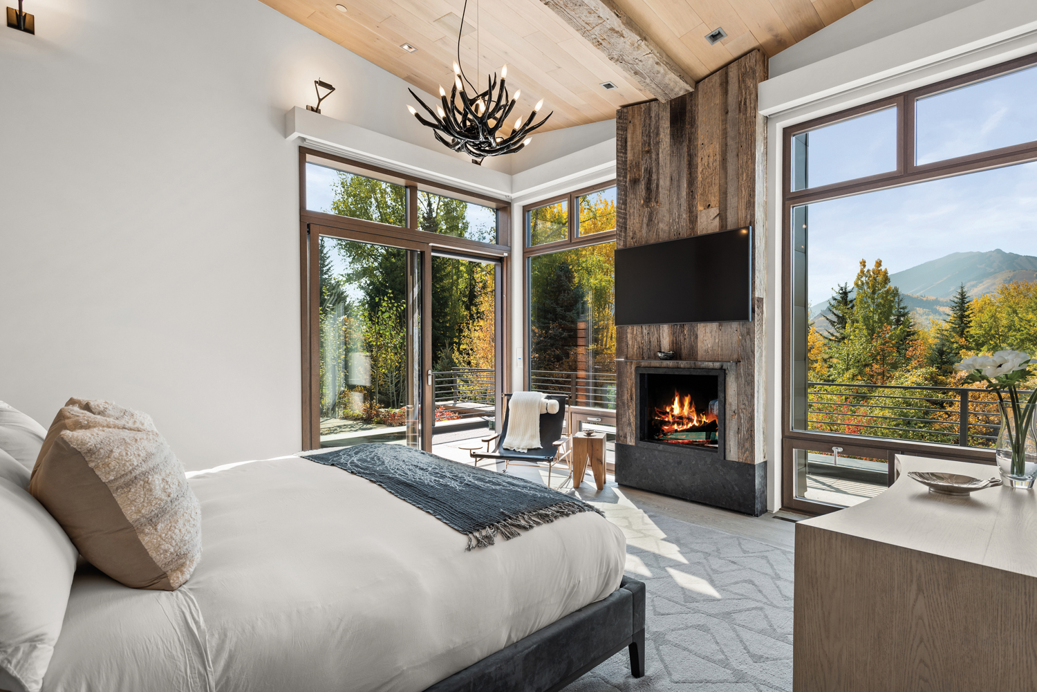 A bedroom designed by Kristin Dittmar with large windows, mountain views in the fall, a fireplace and white bed