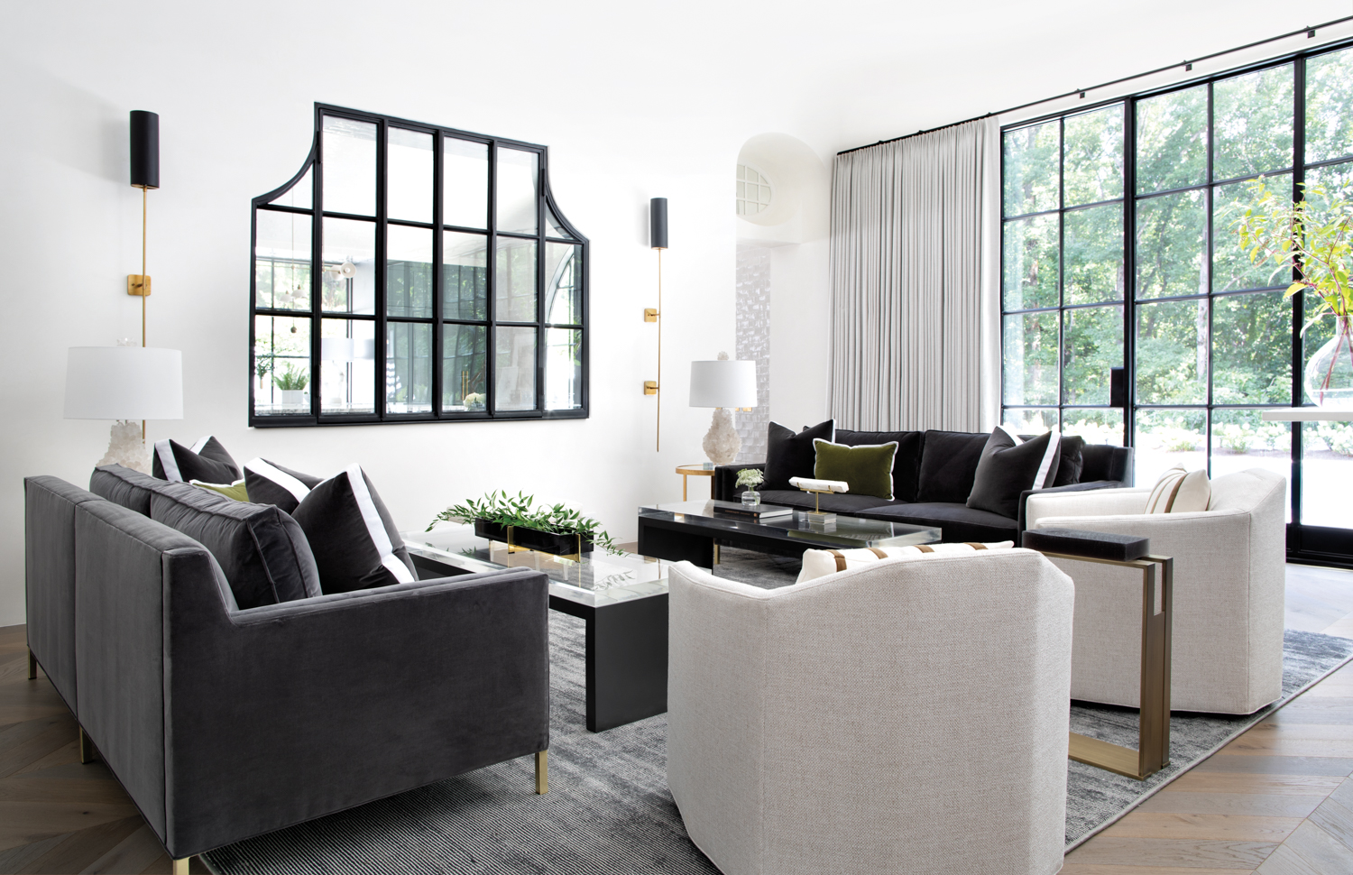 A sunlit living room featuring dark gray sofas, ivory swivel chairs and a large metal window looking into the kitchen