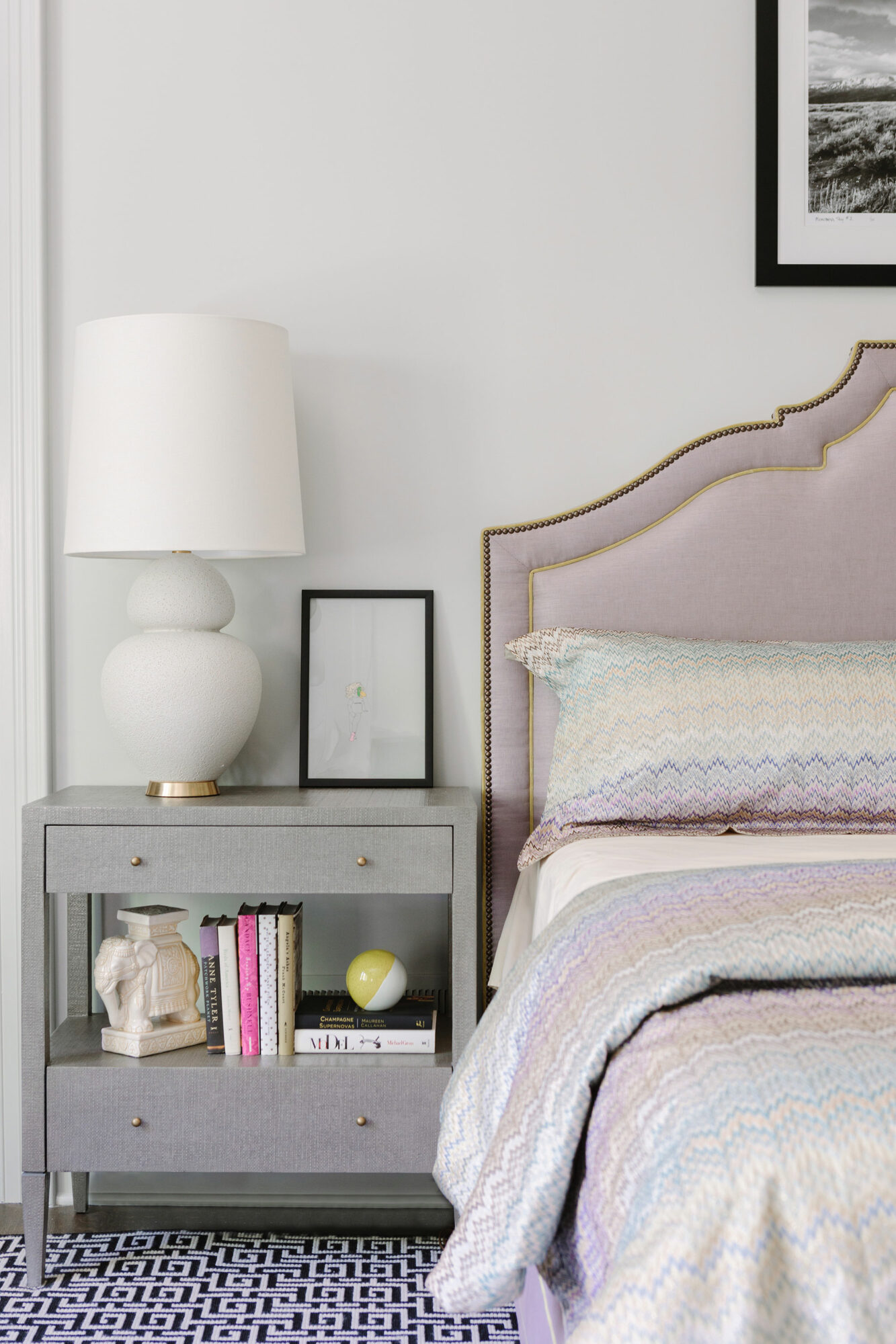 A white bedroom with a lavender upholstered bed dressed in pastel linens.
