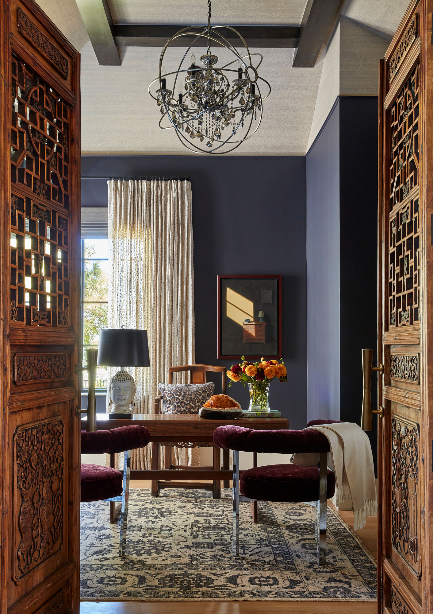 An office with navy walls a wood desk and chair and velvet open-backed guest chairs