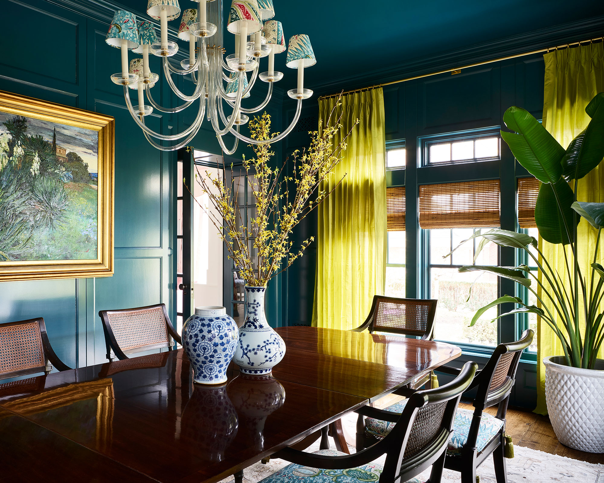 A teal dining room with chartreuse drapes and a a colorful chandelier over a long wood table.