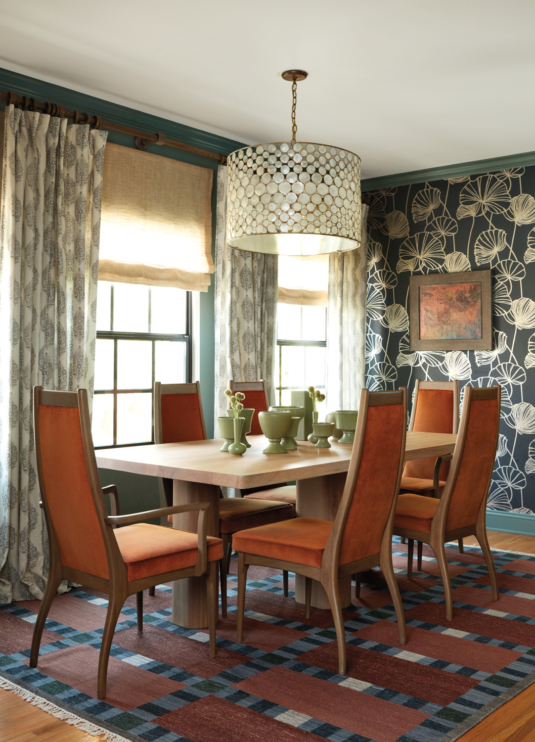 A rectangular, wood dining table circled by tall chairs in an orange fabric, atop a geometric rug and facing an Art Deco-style wallpaper