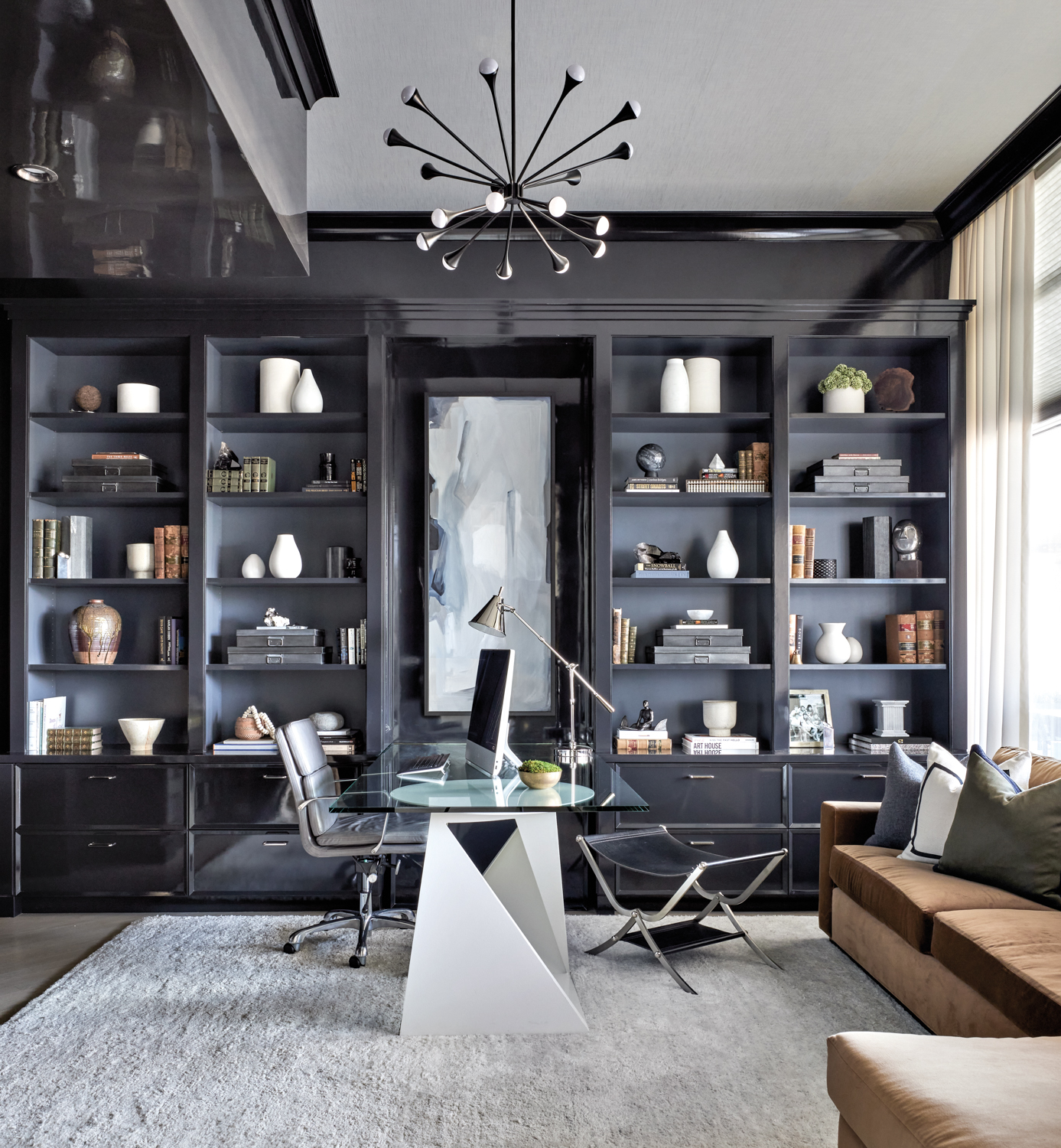 home office with gray shelving, brown couch and a sculptural chandelier above the glass desk