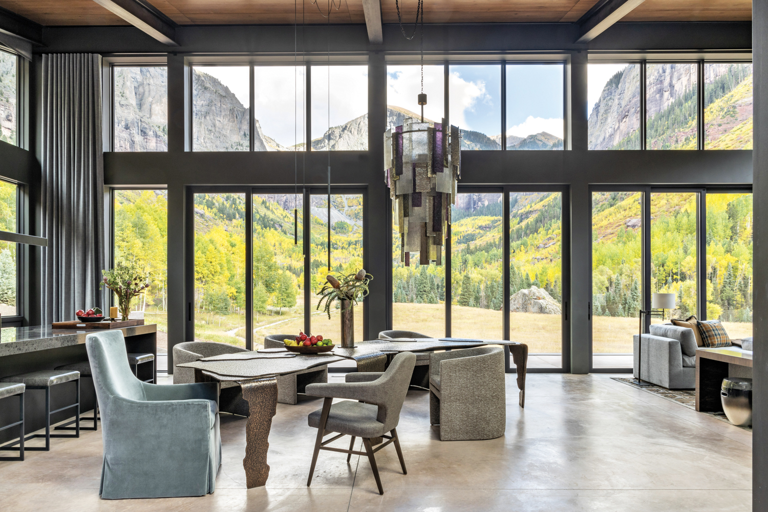 Loft-like dining area with two-story wall of windows, a sculptural chandelier and bronze dining table, varied chairs, and panoramic views outside