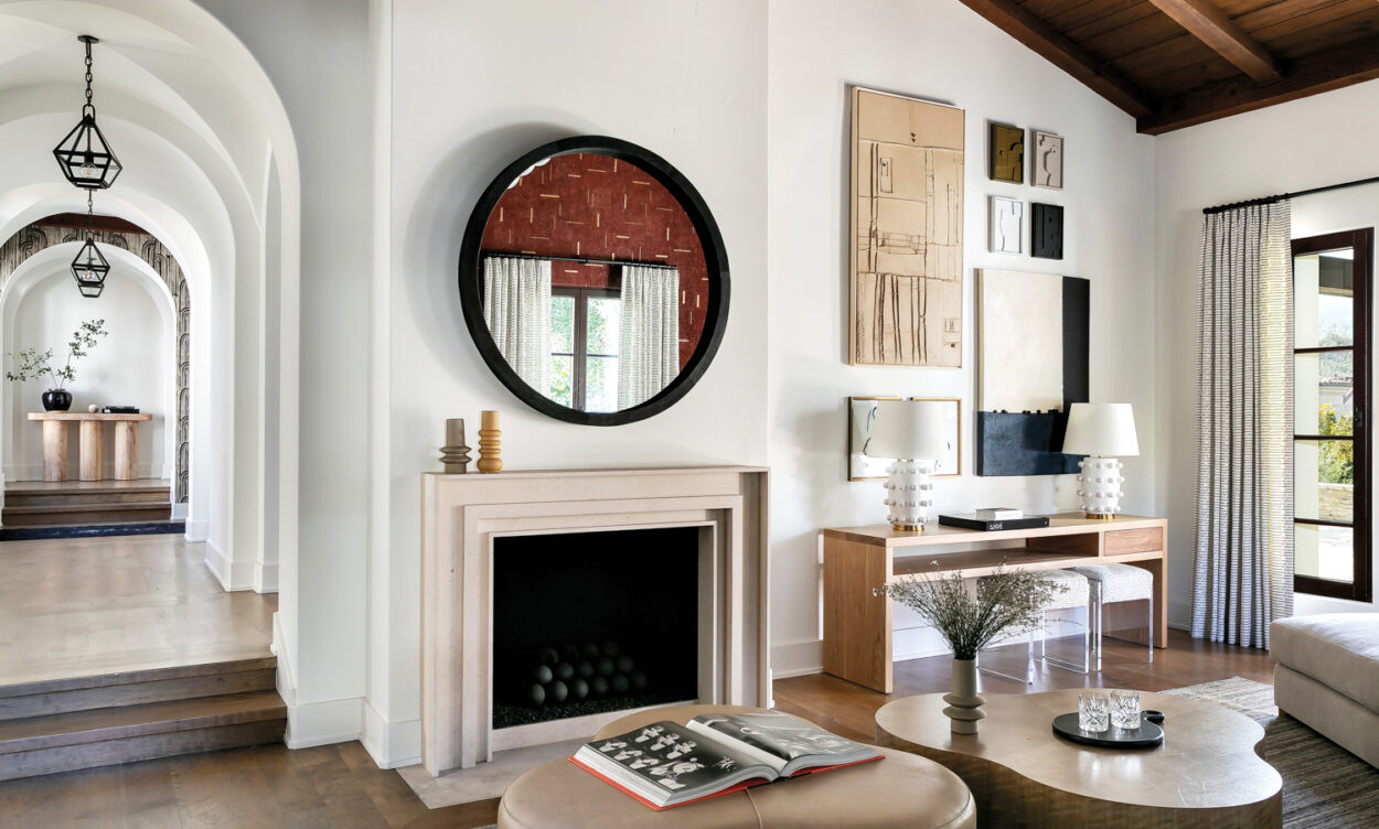 Living room with round, black-framed mirror above fireplace, natural color palette throughout and assortment of abstract paintings