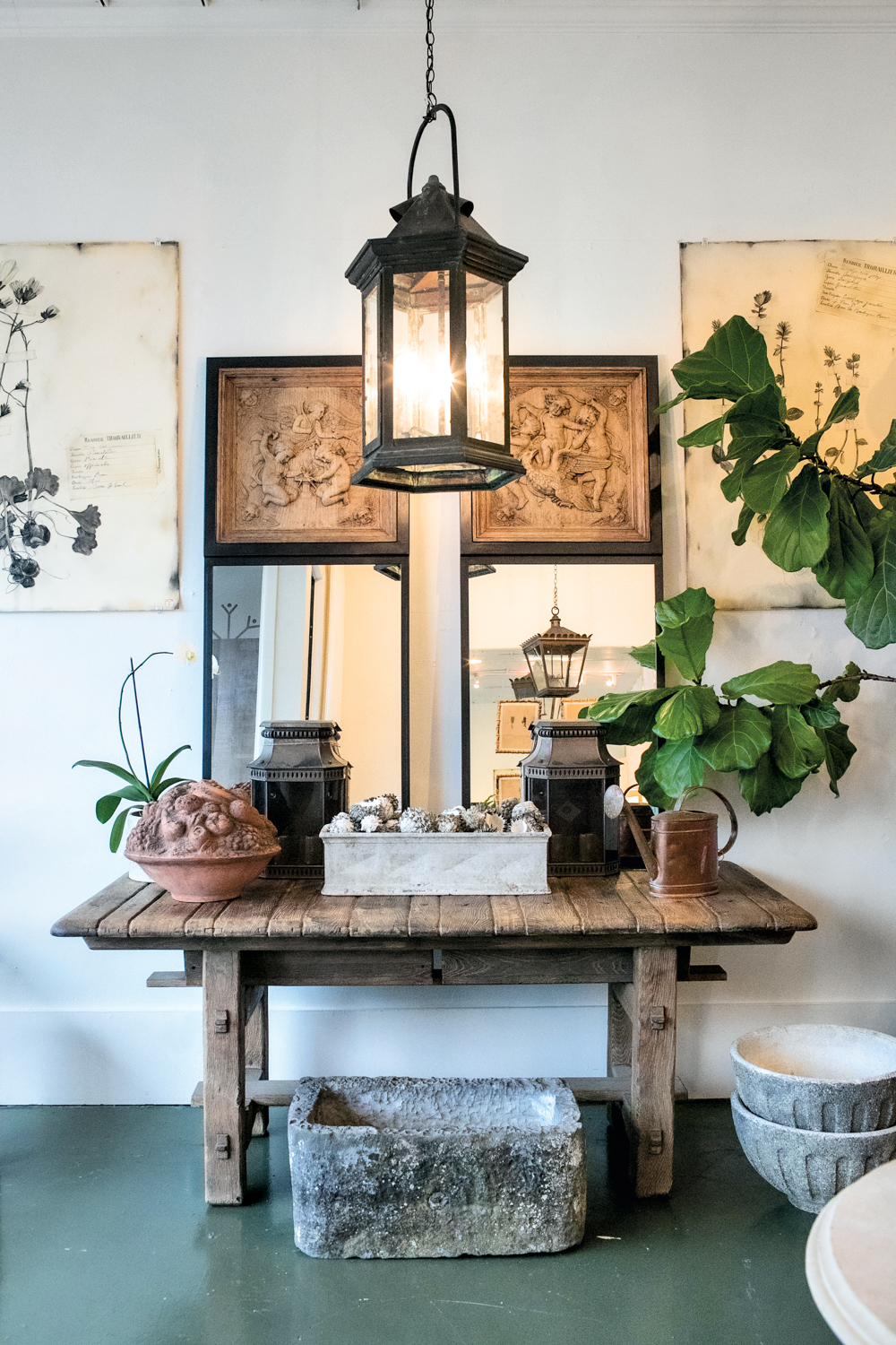 A trestle table holds textural pots and mirrors beneath a lantern, centered between two charcoal floral artworks